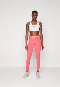 Witte sportbh met zwart Nike-logo, gecombineerd met strakke roze leggings met een witte "Nike Pro" tailleband. Lichte sport schoenen.