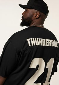 Sort trøje med "THUNDERBOLT" og "21" i hvid skrift. Korte ærmer, rund halsudskæring, mesh tekstur. Bæres med en sort kasket.