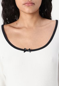 Hvit ribbet topp med svart trim og en liten svart sløyfe ved halslinjen. Har et rundet scoop-design rundt krageområdet.
