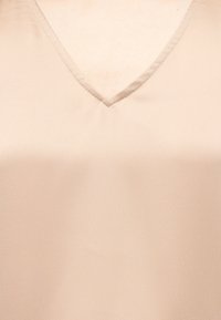 Μπεζ ένδυμα με V-λαιμόκοψη που καλύπτει το πάνω μέρος του στήθους και την περιοχή του λαιμού, με απαλό ύφασμα και διακριτική λεπτομέρεια ραφής.