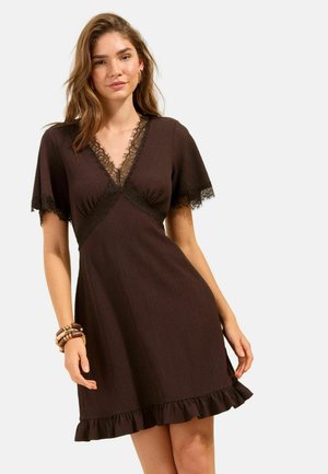 Frau trägt ein kurzes braunes Kleid mit Spitzenbesatz am Ausschnitt und den Ärmeln, gerafftem Saum sowie Armbändern am linken Handgelenk.