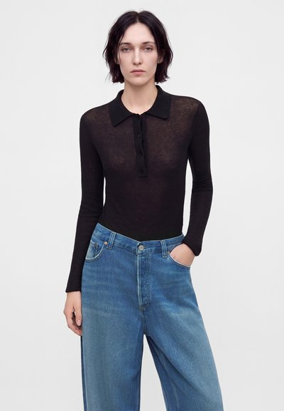 Jeune femme aux cheveux courts et foncés porte un haut noir à manches longues avec col et un jean bleu ample avec une main dans la poche.