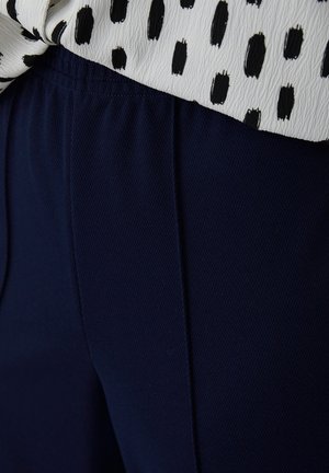 Pantalones de tela texturizada azul marino con cintura elástica, combinados con una camisa blanca con un patrón de puntos negros. Vista en primer plano.