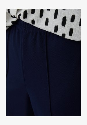 Marineblauwe textuurstof broek met een elastische tailleband, gecombineerd met een wit shirt met een zwart stippenpatroon. Close-up weergave.