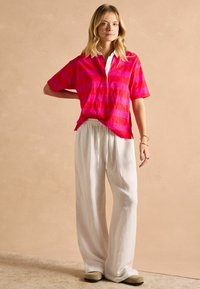 Ružový pruhovaný polo tričko s krátkymi rukávmi a bielym golierom, spárované s voľnými bielymi nohavicami a béžovými šľapkami. Svetlé pozadie.