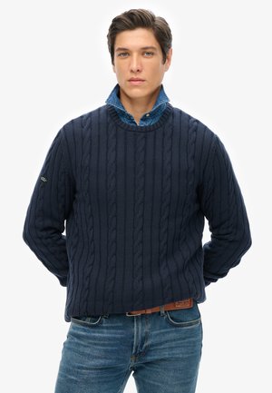 Jersey de punto trenzado en color azul marino con un diseño de cuello redondo, que presenta detalles texturizados, y se lleva encima de una camisa de denim azul claro.