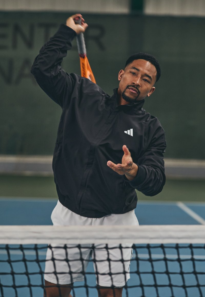 Zwarte sportjack met prominent drie-stripes logo, witte shorts, speler in actie die zich voorbereidt om een tennisbal te slaan, binnenveldsetting.