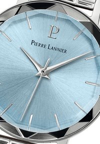 Reloj de metal plateado con una esfera texturada de color azul claro, manecillas de hora y minuto plateadas, y acentos en forma de triángulo en negro alrededor del bisel. Marca: Pierre Lannier.