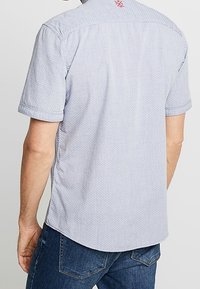 Camisa de manga corta en tela de rayas azul claro y blanca. Presenta un dobladillo curvado y un logo rojo en el cuello trasero. Corte casual.
