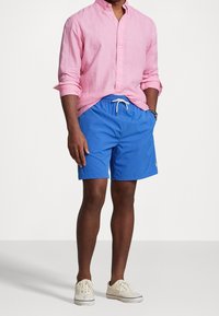 Ljusrosa linne skjorta med knapp-ner krage, ihop med blå badshorts med elastisk midja och justerbar dragsko. Vita sneakers.