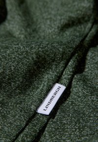Ciemnozielona tkanina o teksturze z delikatnym wzorem, z białą metką oznaczoną "LINDHBERGH" wszytą w szew.
