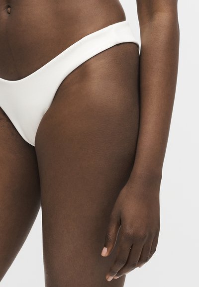 Gros plan d'une personne à la peau foncée portant un bas de bikini blanc, mettant en valeur sa hanche, sa cuisse supérieure et sa main détendue sur le côté.