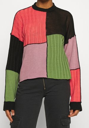Femme portant un pull côtelé à blocs de couleurs avec des patches roses, noirs et verts, associé à un pantalon cargo noir.