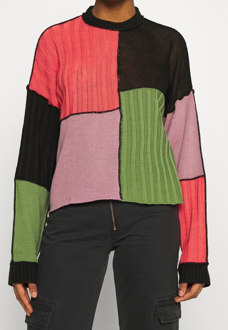 Femme portant un pull côtelé à blocs de couleurs avec des patches roses, noirs et verts, associé à un pantalon cargo noir.