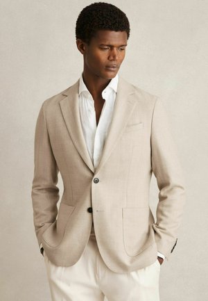 Beige Leinenblazer mit einreihigem Knopfverschluss und zwei Fronttaschen; kombiniert mit einem hellen Hemd und cremefarbenen Hosen. Glatte Textur.