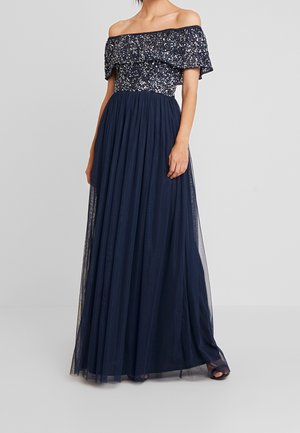 Femme vêtue d'une robe de soirée bleu marine sans épaules avec un corsage à paillettes et une jupe en tulle fluide, se tenant devant un fond uni.