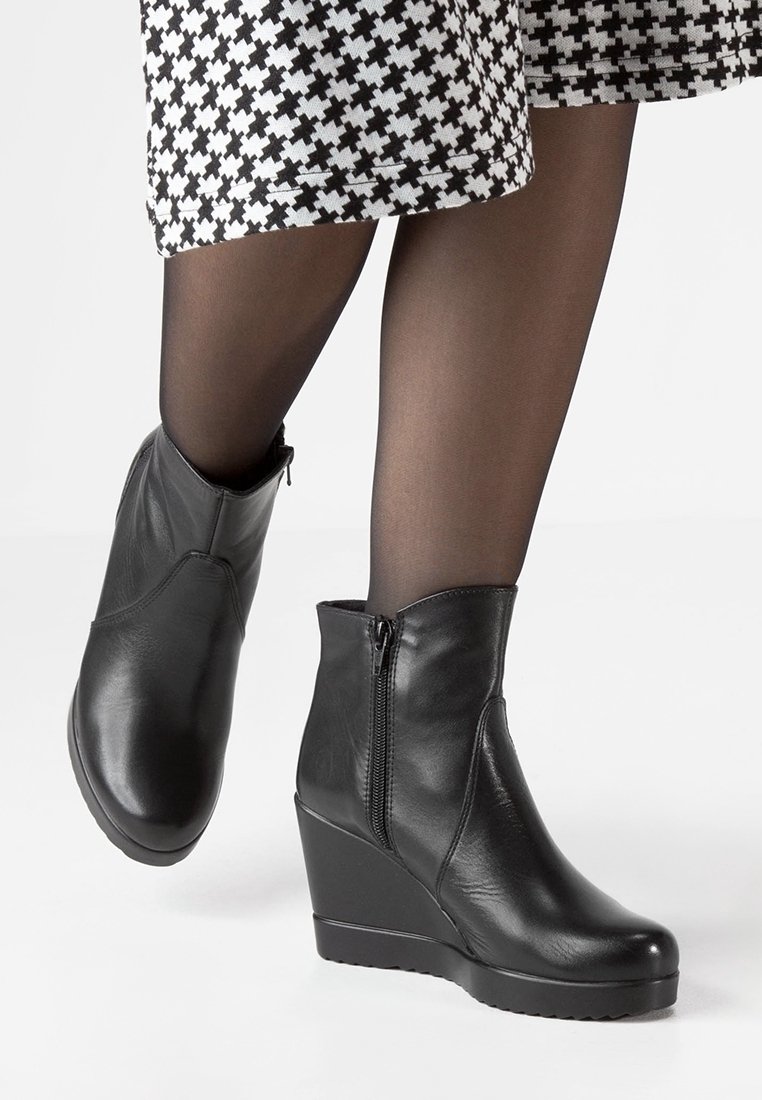 Bottines en cuir noir à talon compensé, avec fermeture éclair sur le côté, texture lisse et bout arrondi. Illustrées avec une jupe à motifs et des collants sombres.