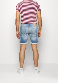 Shorts in denim azzurro con risvolti, caratterizzati da due tasche posteriori e una patch in pelle marrone sopra la vita. Camicia a righe rossa e bianca visibile.