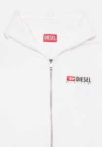 Veste à zip blanche faite de tissu doux, avec un logo "DIESEL" rouge et noir sur la poitrine gauche. Comprend une capuche et des poches avant.