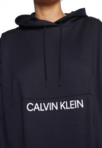 Felpa nera con cappuccio e lacci, caratterizzata da un logo bianco "CALVIN KLEIN" sul davanti. Tessuto morbido con una texture liscia.