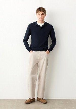 Mladić stoji, nosi tamnoplavu polo majicu s dugim rukavima, bijelu majicu ispod, svijetlo bež hlače i smeđe casual cipele ispred jedinstvenog bijelog zida.