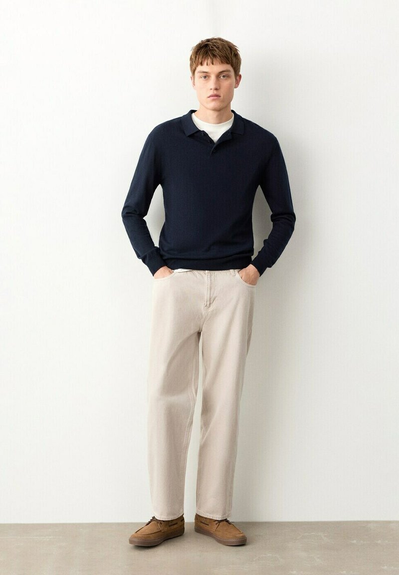 Mladý muž stojí, oblečený v tmavomodrom polo tričku s dlhým rukávom, bielom tričku pod ním, svetlo béžových nohaviciach a hnedých pohodlných topánkach, na pozadí bielej steny.