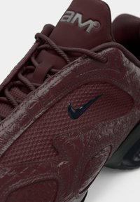 Burgundske teniske copate z teksturirano zgornjšo stranjo, mrežastim materialom, izstopajočim črnim Nike znakom, podloženim podplatom ter debelimi vezalkami.