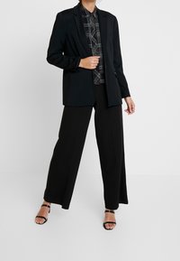 Schwarzer Blazer mit Revers, kombiniert mit einem karierten Hemd mit Knöpfen und weiten schwarzen Hosen. Schwarze Sandalen mit offener Zehe.