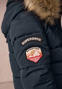 Chaqueta de plumas azul marina con capucha de piel sintética. Presenta un parche bordado que dice "SUPERDRY" y "EXPEDITION EVEREST" con gráficos de montañas.