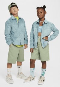 Du vaikai pozuoja derančiais drabužiais. Berniukas vilki šviesiai mėlyną aksominį marškinį, žalias šortus ir sportbačius. Mergaitė dėvi mėlyną trumpą viršų, žalias šortus ir spalvotus kojines.