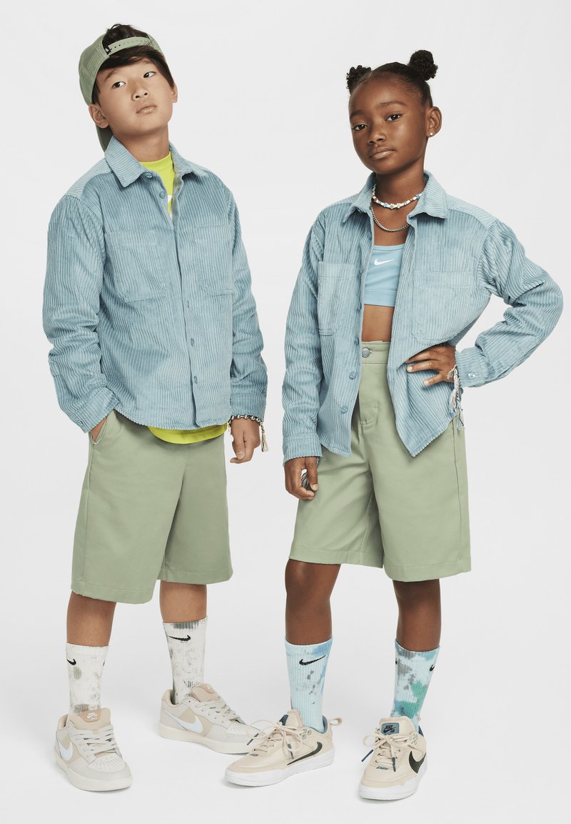 Du vaikai pozuoja derančiais drabužiais. Berniukas vilki šviesiai mėlyną aksominį marškinį, žalias šortus ir sportbačius. Mergaitė dėvi mėlyną trumpą viršų, žalias šortus ir spalvotus kojines.