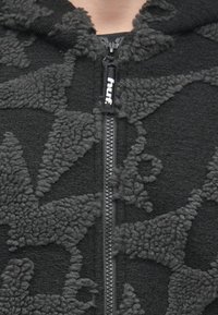 Melna vilnas vējjakas ar mīkstu tekstūru, kurai ir kontrastējošs pelēks raksts un priekšējā rāvējslēdzēja ar zīmola cilpiņu.