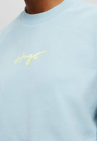 Sudadera azul clara con cuello acanalado, que presenta un logotipo bordado en amarillo "Hugo" en el pecho. Textura de tela suave.