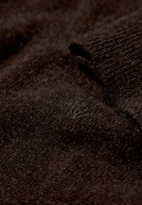 Braun gestrickter Pulloverstoff mit weicher Textur, der ein dezentes gesticktes Emblem aufweist. Eine Nahaufnahme betont das kunstvolle Gewebe.