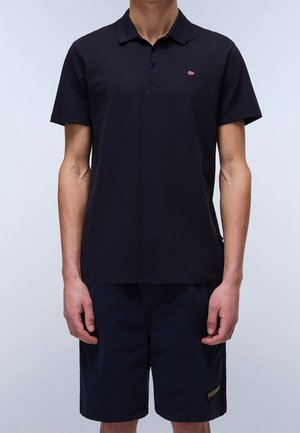 Mand iført en sort kortærmet poloshirt med et lille logo på brystet og sorte shorts mod en lys baggrund.