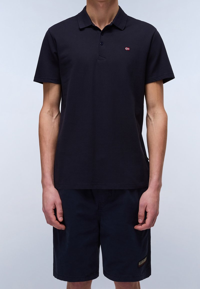 Homme portant un polo noir à manches courtes avec un petit logo sur la poitrine et un short noir sur un fond clair.