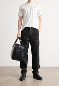 Bílý bavlněný tričko s malým černým logem, spárované s tmavými džínovými kalhotami, držící černou koženou tašku s dvojitými rukojetmi. Černé boty.