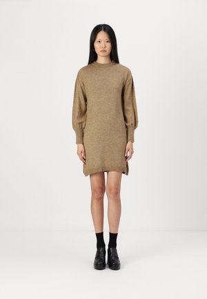 Knit sweater dress in light brown, featuring a high neckline, puffed long sleeves, and side slits at the hem. Black footwear complements the look.