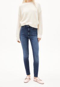 Pull côtelé crème, jean skinny taille haute bleu foncé et chaussures rose pâle avec détail à bride. Texture douce et coupe ajustée.