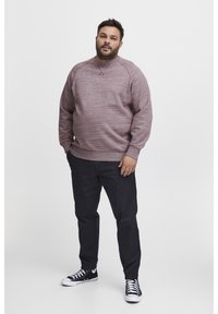 Man met baard, gekleed in een bordeauxrode sweatshirt, zwarte joggingsbroek en zwarte sneakers, staande tegen een effen witte achtergrond.