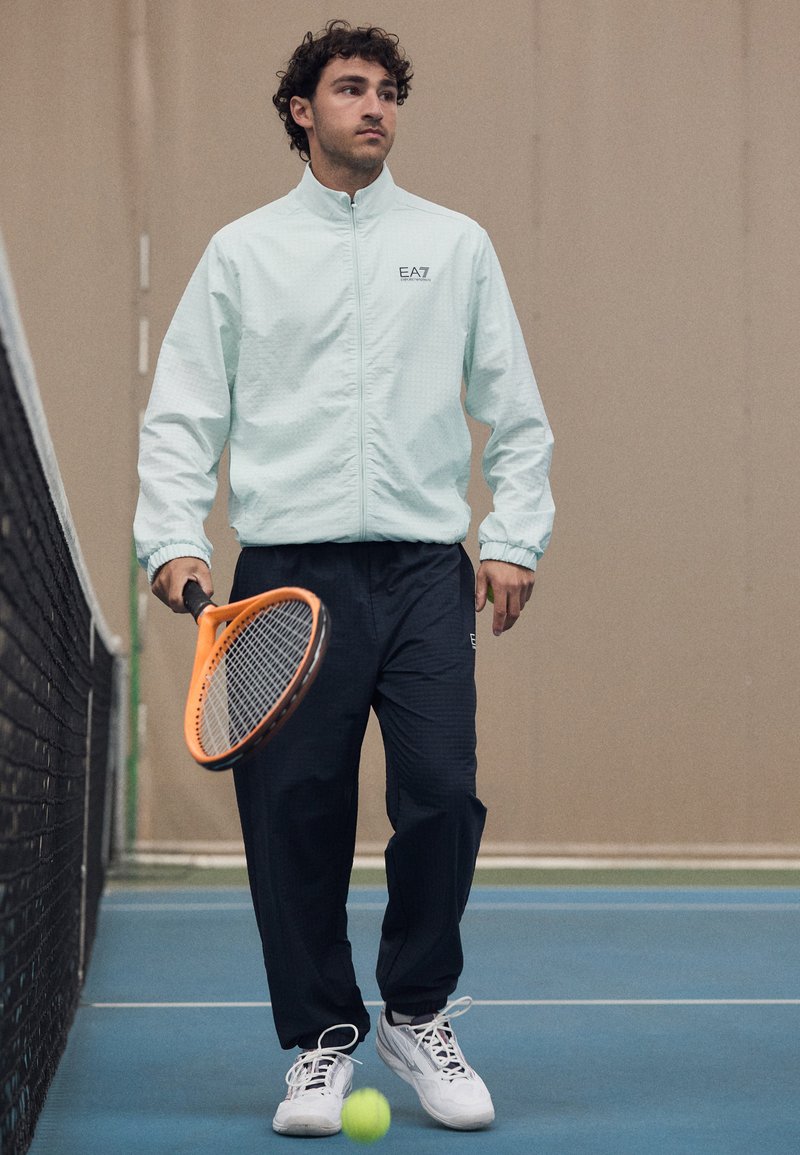 Casaco azul-claro com fecho, padrão texturizado, calças azul-marinho e tênis brancos. Segurando uma raquete de ténis laranja e de pé numa quadra.