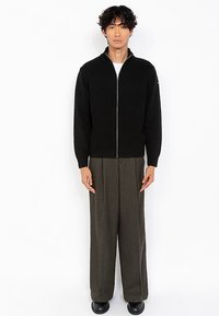 Zwarte geribbelde gebreide cardigan met rits en hoge kraag, gedragen over grijze broek met wijde pijpen, tegen een eenvoudige witte achtergrond.