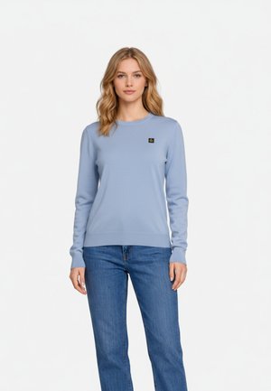 Kobieta z falującymi blond włosami, ubrana w jasnoniebieski sweter z logo na piersi oraz niebieskie dżinsy, stojąca na tle białego tła.