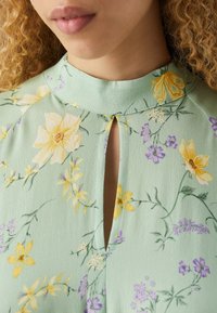 Grünes Blumenkleid mit hohem Kragen, das mit gelben und lila Blumen verziert ist, über ein Schlüsselloch-Detail verfügt und aus leichtem, strukturiertem Stoff besteht.