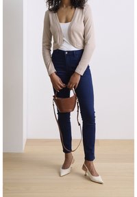 Beiger, kurzer Cardigan über einem weißen Tanktop, kombiniert mit dunkelblauen Skinny Jeans und weißen Slingback-Pumps. Hält eine kleine braune Handtasche.