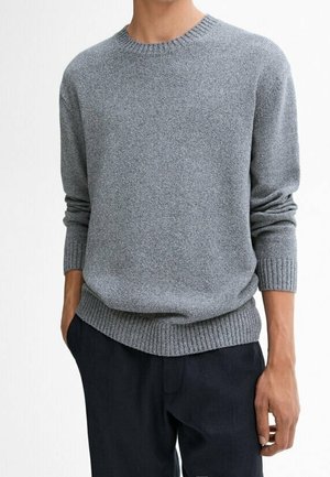 Homme portant un pull gris uni à col rond avec poignets et base côtelés, associé à un pantalon foncé sur un fond neutre.