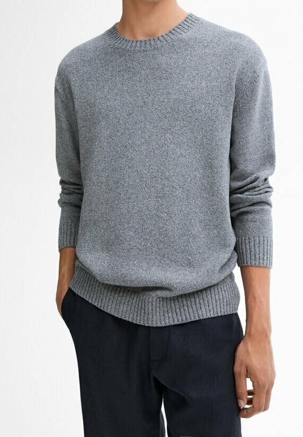 Homme portant un pull gris uni à col rond avec poignets et base côtelés, associé à un pantalon foncé sur un fond neutre.