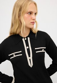 Maglietta nera a maniche lunghe con colletto a punta, dettagli color crema, due tasche sul petto con righe orizzontali e chiusura a bottoni.
