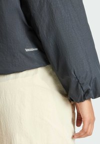 Detailný záber na ruku opretú o pás, ktorá má na sebe tmavosivú bundu climawarm a svetlobežovú textúrovanú sukňu alebo nohavice.