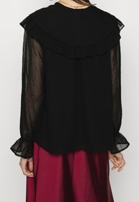 Blusa nera con colletto arricciato e maniche lunghe trasparenti. Il materiale strutturato sembra leggero. Vista da dietro, abbinata a una gonna bordeaux.